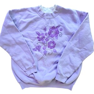 Puede incluir: Una sudadera de color lavanda con un diseño floral en tonos morados. Las flores están bordadas con pedrería.
