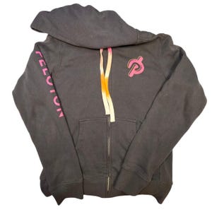 May include: A charcoal gray zip-up hoodie with a hood and a front pocket. The word "PELOTON" is printed in pink on the left sleeve. The Peloton logo is on the left chest. The drawstring is pink and orange.