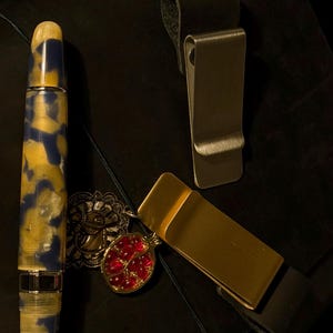 May include: A fountain pen with a blue and yellow marbled design, a gold money clip, and a silver money clip are displayed on a dark surface. A red and gold pendant and a gold-colored decorative element are also visible.