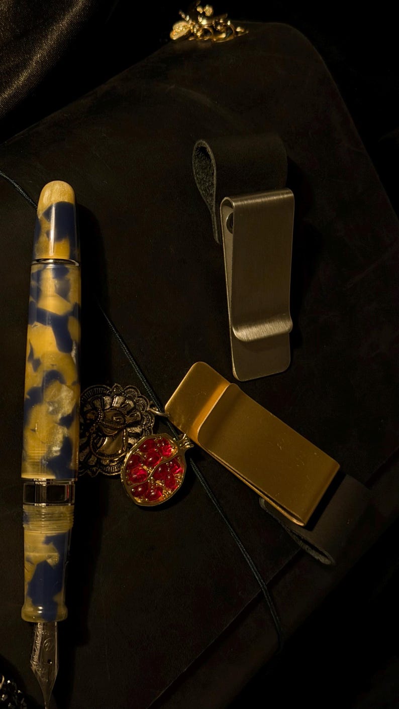 May include: A fountain pen with a blue and yellow marbled design, a gold money clip, and a silver money clip are displayed on a dark surface. A red and gold pendant and a gold-colored decorative element are also visible.