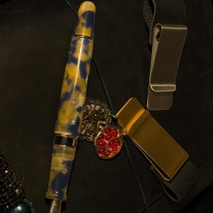 May include: A collection of luxury writing accessories, including a fountain pen with a blue and yellow marbled design, a gold money clip, and a silver money clip. A red jeweled pendant and a beaded pen holder are also present.