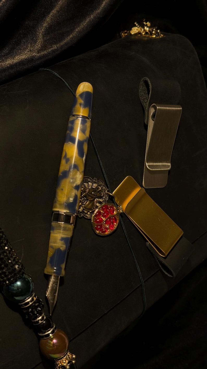 May include: A collection of luxury writing accessories, including a fountain pen with a blue and yellow marbled design, a gold money clip, and a silver money clip. A red jeweled pendant and a beaded pen holder are also present.