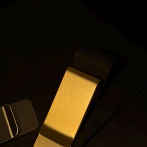 May include: A gold-colored money clip with a decorative pendant featuring a red gemstone design. The clip is rectangular with a curved edge. The pendant is attached to the clip with a small chain. The background is dark.