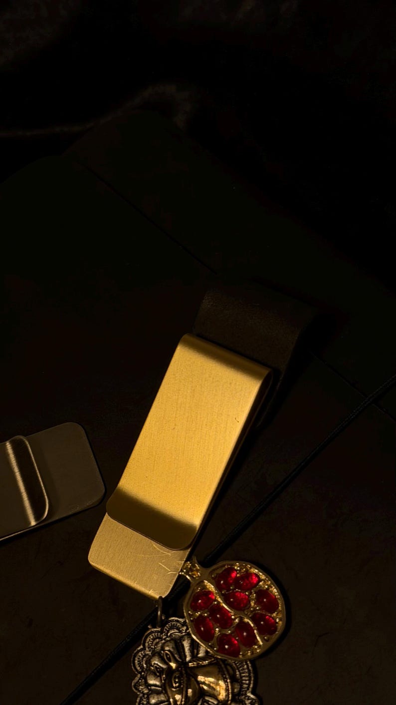 May include: A gold-colored money clip with a decorative pendant featuring a red gemstone design. The clip is rectangular with a curved edge. The pendant is attached to the clip with a small chain. The background is dark.