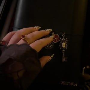 May include: Black leather bag with decorative charms. Charms include a key, a cameo, and a red jeweled pendant. The bag is held by a hand with long, pointed nails with a black and white design. A gold ring with a red stone is also visible.
