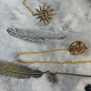 Puede incluir: Una colección de marcapáginas decorativos de metal con varios amuletos. Los marcapáginas presentan diseños de plumas en tonos plateados y bronce, junto con amuletos como un sol, un dragón, una mano de Hamsa y un ojo turco. Los marcapáginas están conectados a cadenas.