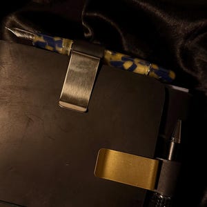 May include: A close-up of a black notebook with a pen and pen holder. The pen has a blue and yellow marbled design. Two metal pen holders are attached to the notebook. Two decorative charms hang from the notebook.