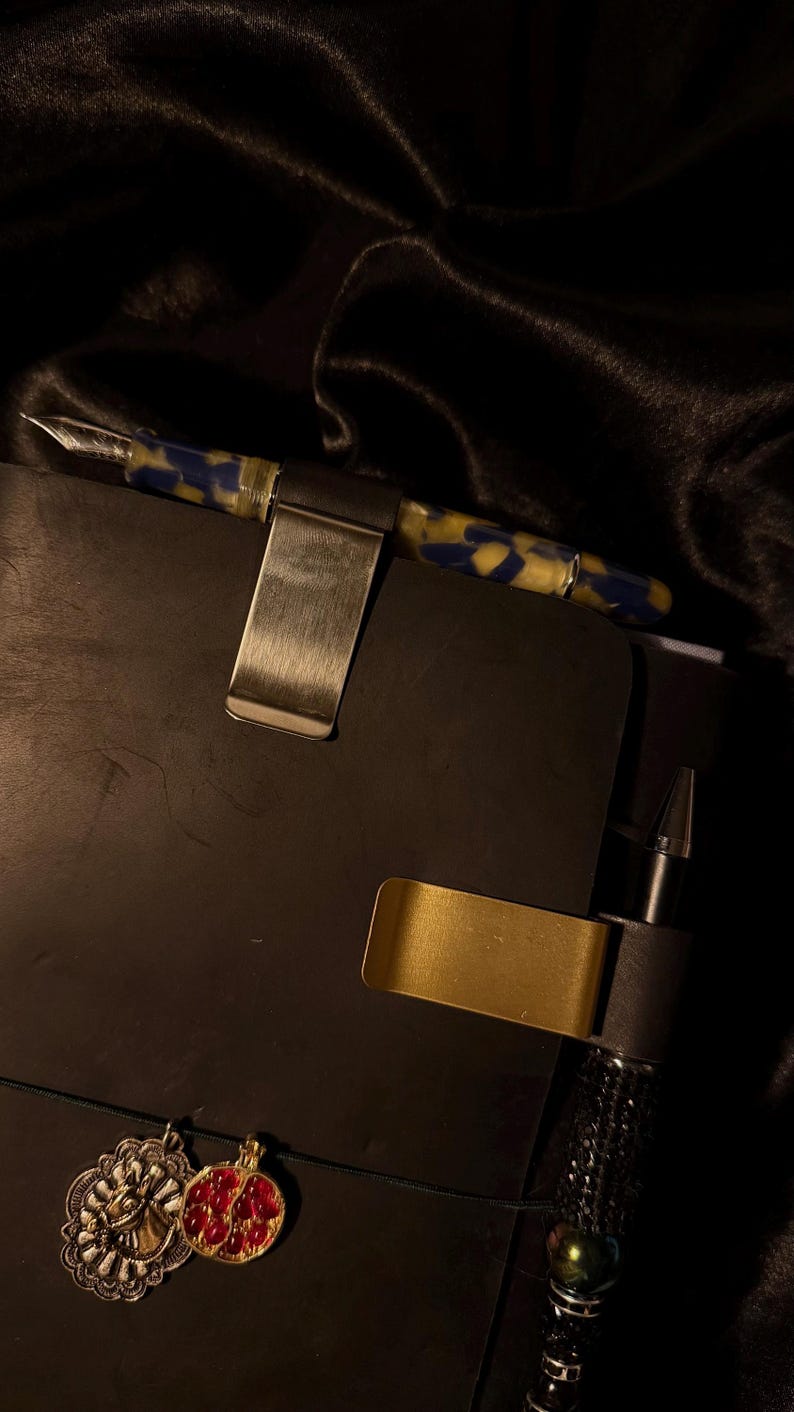 May include: A close-up of a black notebook with a pen and pen holder. The pen has a blue and yellow marbled design. Two metal pen holders are attached to the notebook. Two decorative charms hang from the notebook.
