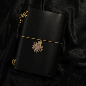 May include: Black leather journal with a gold-colored clasp and a decorative charm. The charm features a pink gemstone in a gold filigree setting. The journal is closed with a gold elastic band.