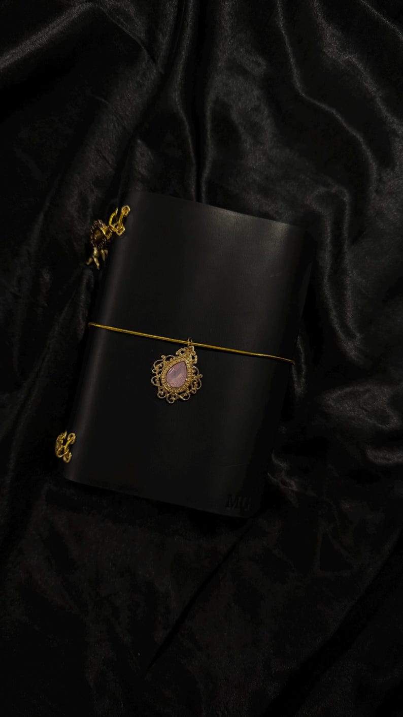 May include: Black leather journal with a gold-colored clasp and a decorative charm. The charm features a pink gemstone in a gold filigree setting. The journal is closed with a gold elastic band.