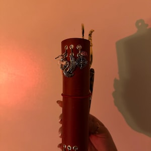 May include: A cylindrical, burgundy-colored leather journal with decorative charms. Charms include a swan, a compass, and keys. The journal is held by a hand, and the background is a warm, peachy color.