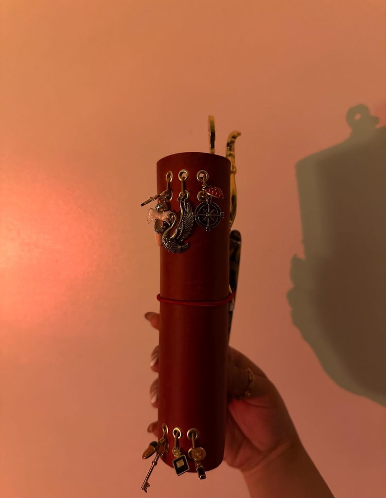 May include: A cylindrical, burgundy-colored leather journal with decorative charms. Charms include a swan, a compass, and keys. The journal is held by a hand, and the background is a warm, peachy color.