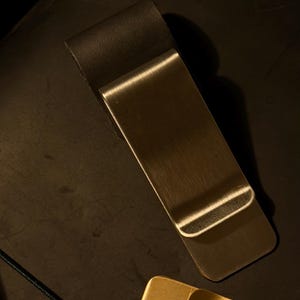 May include: A silver-colored metal money clip with a curved top and a rectangular body. The clip is lying on a dark surface, with a gold-colored money clip and jewelry in the background.