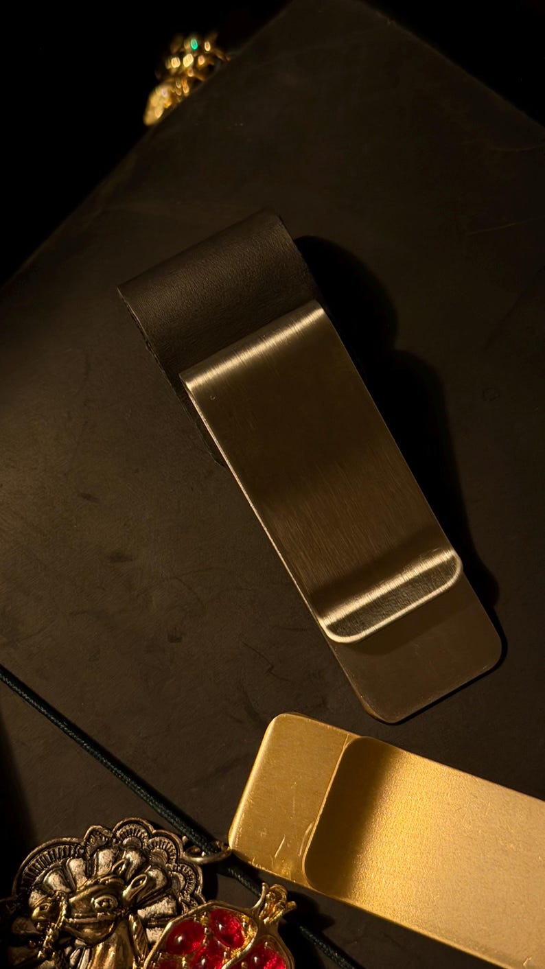 May include: A silver-colored metal money clip with a curved top and a rectangular body. The clip is lying on a dark surface, with a gold-colored money clip and jewelry in the background.