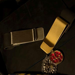 May include: Two metal money clips, one silver and one gold, rest on a dark surface. The gold clip has a decorative charm with a red gem design. A pen with a blue and yellow pattern is also visible.