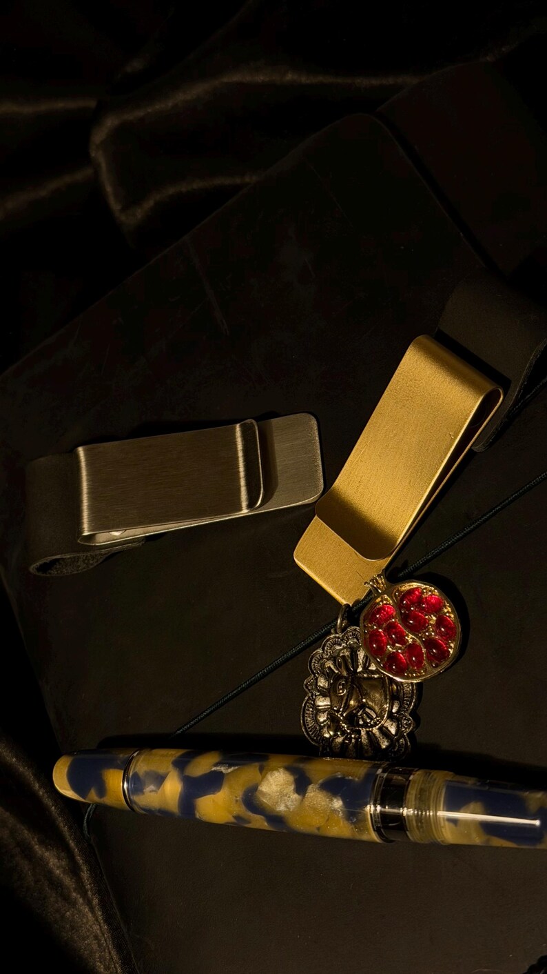 May include: Two metal money clips, one silver and one gold, rest on a dark surface. The gold clip has a decorative charm with a red gem design. A pen with a blue and yellow pattern is also visible.