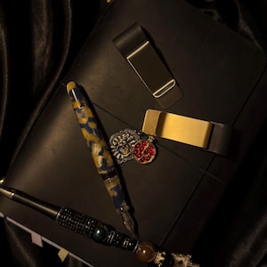 Puede incluir: Un diario de cuero negro con un bolígrafo dorado, un bolígrafo con pedrería negra y dos clips para billetes. También hay un bolígrafo con un patrón azul y amarillo y un colgante rojo y dorado. Los artículos están dispuestos sobre una superficie de satén negro.