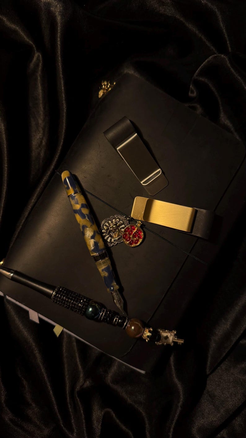May include: A black leather journal with a gold-colored pen, a pen with black rhinestones, and two money clips. A pen with a blue and yellow pattern and a red and gold pendant are also present. The items are arranged on a black satin surface.