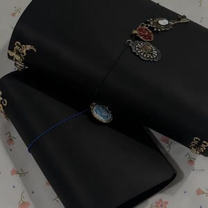 May include: Two black leather journals with decorative charms attached. One journal has a blue elastic closure. The charms are various shapes and colors, including gold, red, and white. The journals are on a floral patterned surface.