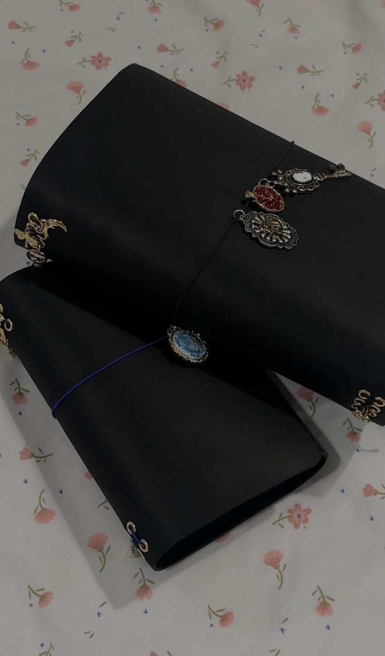 May include: Two black leather journals with decorative charms attached. One journal has a blue elastic closure. The charms are various shapes and colors, including gold, red, and white. The journals are on a floral patterned surface.