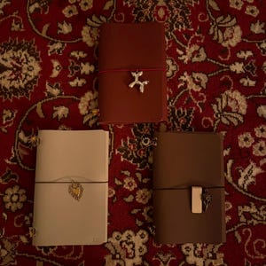 May include: Three leather journals in shades of brown and cream, each with a unique charm. The journals are closed with an elastic band. One journal has a dog charm, another a heart, and the third a decorative charm.