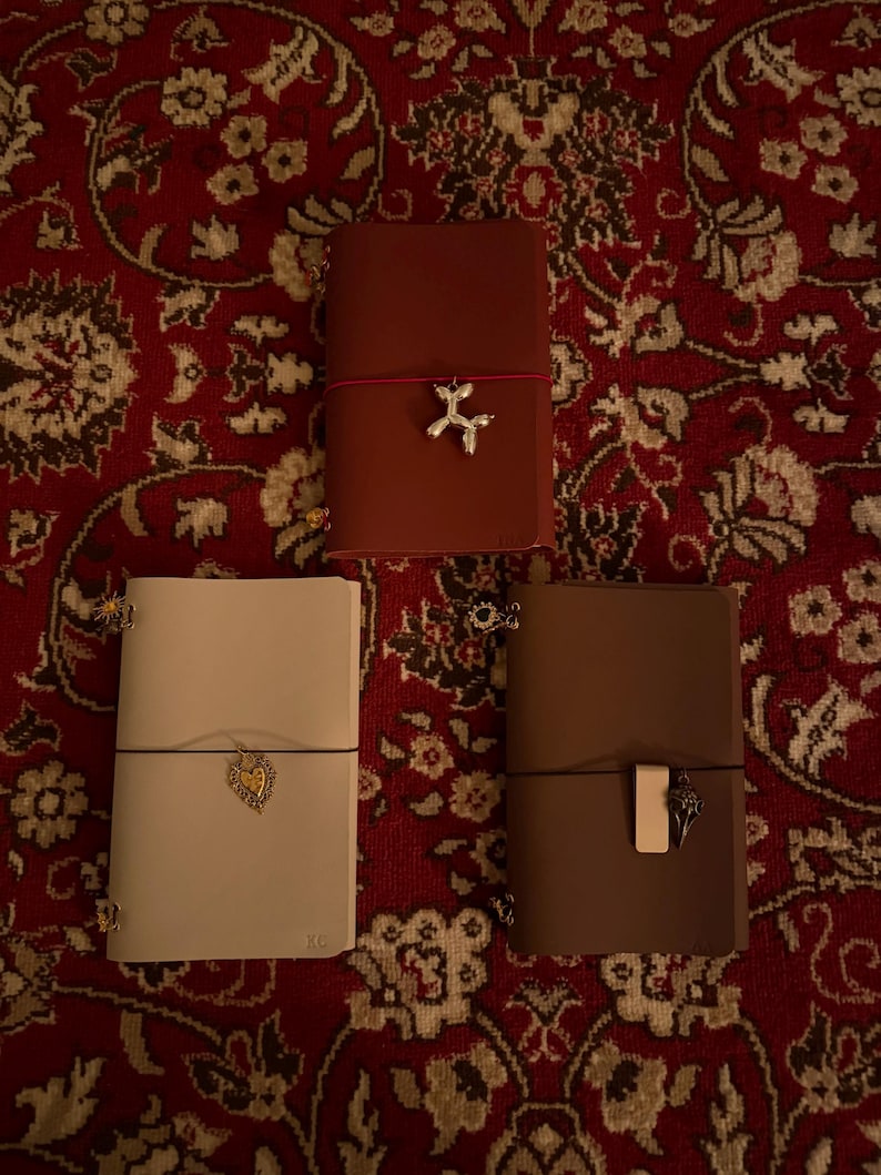 May include: Three leather journals in shades of brown and cream, each with a unique charm. The journals are closed with an elastic band. One journal has a dog charm, another a heart, and the third a decorative charm.