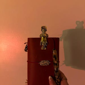 May include: A burgundy leather journal with a gold-colored hand-shaped clip and a pen with a speckled pattern. The journal is secured with a red elastic band and a small decorative charm. A small bracelet with a speckled stone is also visible.