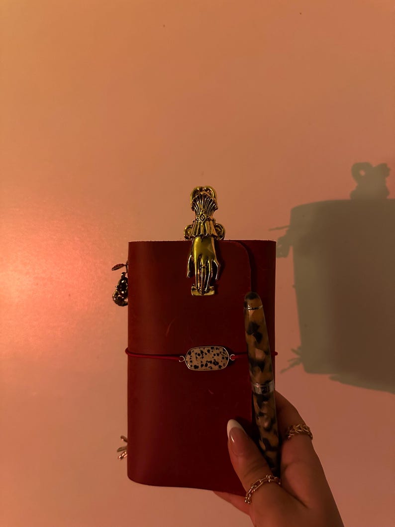 May include: A burgundy leather journal with a gold-colored hand-shaped clip and a pen with a speckled pattern. The journal is secured with a red elastic band and a small decorative charm. A small bracelet with a speckled stone is also visible.