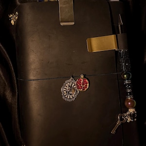 May include: A black leather journal with a metal clip and gold accents. The journal has a pen holder with a beaded charm and decorative charms. A pen with a blue and yellow pattern is clipped to the top.
