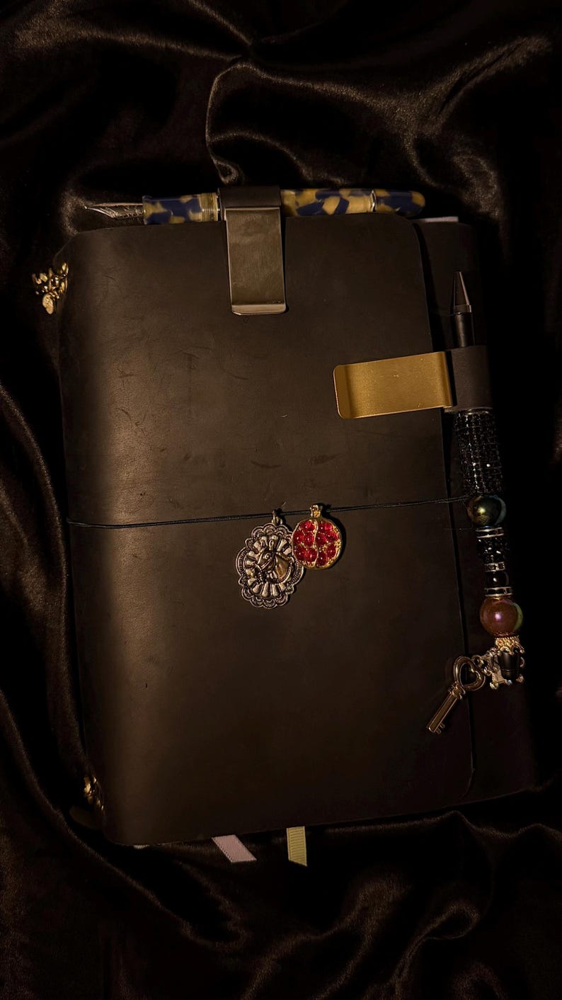 May include: A black leather journal with a metal clip and gold accents. The journal has a pen holder with a beaded charm and decorative charms. A pen with a blue and yellow pattern is clipped to the top.