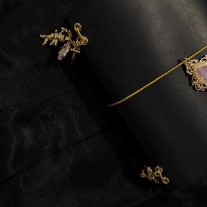 May include: A black leather journal with a gold elastic closure and decorative gold charms. The journal has a gold charm with a pink stone and a gold charm with a small angel. The background is a black fabric.