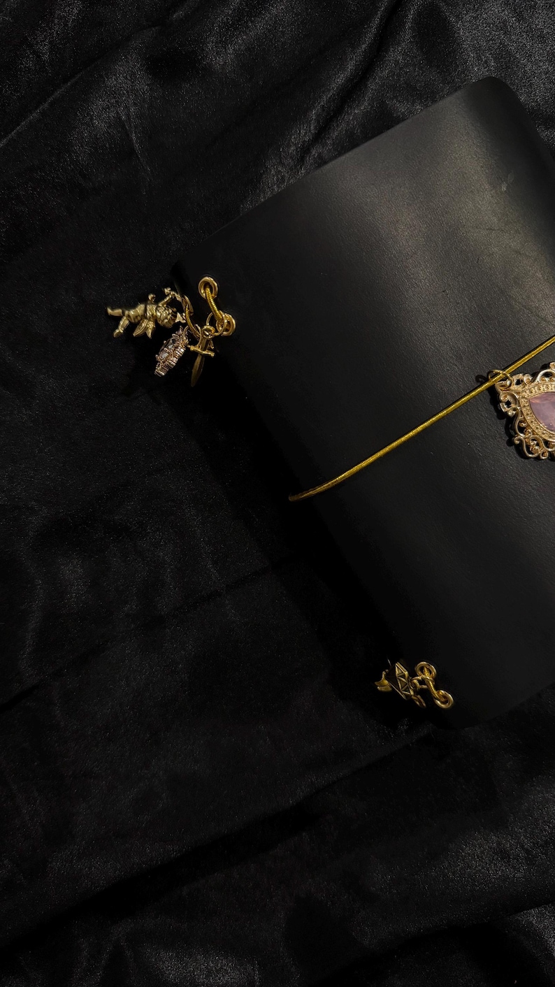 May include: A black leather journal with a gold elastic closure and decorative gold charms. The journal has a gold charm with a pink stone and a gold charm with a small angel. The background is a black fabric.