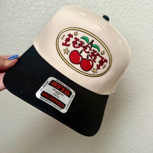 May include: A beige and black baseball cap with the word "Lucky" embroidered in red and gold, along with a pair of cherries. The cap has a curved brim and a tag that reads "OTTO Classic Fit".