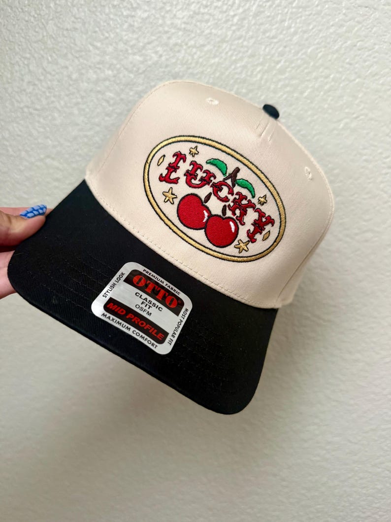 May include: A beige and black baseball cap with the word "Lucky" embroidered in red and gold, along with a pair of cherries. The cap has a curved brim and a tag that reads "OTTO Classic Fit".