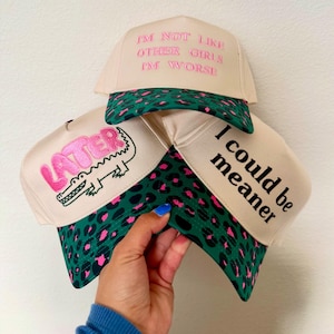 May include: Three beige trucker hats with green and pink leopard print brims. One hat features pink embroidered text reading I'M NOT LIKE OTHER GIRLS I'M WORSE. Another has a pink embroidered alligator and the word LATER. The third has black text I could be meaner.