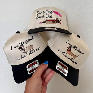 May include: Three cream-colored baseball caps with black brims. The top cap features a hot dog graphic and the text "Suns Out Buns Out." The two lower caps feature dog illustrations wearing cowboy hats and the text "I am the friend in low places."