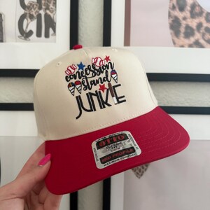 May include: A baseball cap with a cream-colored crown and a red brim. The cap features the embroidered text "Concession Stand Junkie" with baseball, star, and ice cream cone graphics. The cap has a classic fit and a mid profile.