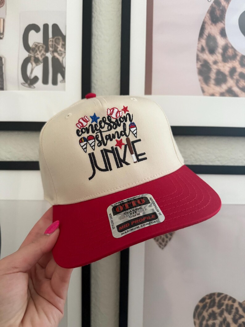 May include: A baseball cap with a cream-colored crown and a red brim. The cap features the embroidered text "Concession Stand Junkie" with baseball, star, and ice cream cone graphics. The cap has a classic fit and a mid profile.