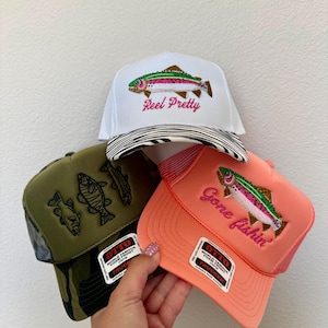 May include: Three trucker hats with fishing-themed designs. One white hat with a rainbow trout and the text "Reel Pretty." Another is coral with "Gone fishin'" and a trout. The third is olive green with fish graphics.