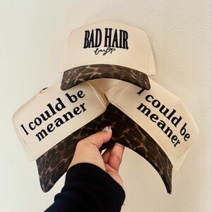 May include: Three cream-colored trucker hats with brown leopard print brims. The top hat features the embroidered text "BAD HAIR day" in black. The two lower hats feature the black printed text "I could be meaner" on the front panels.