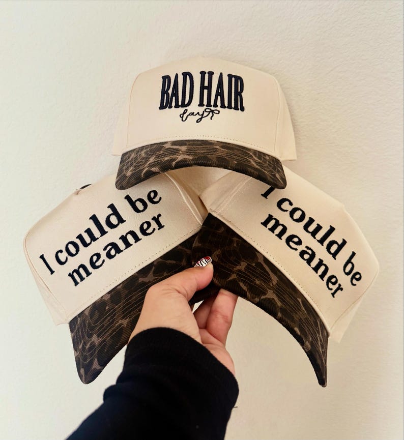 May include: Three cream-colored trucker hats with brown leopard print brims. The top hat features the embroidered text "BAD HAIR day" in black. The two lower hats feature the black printed text "I could be meaner" on the front panels.