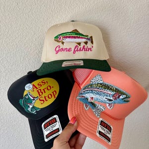 May include: Three trucker hats with embroidered fish designs. One hat is tan with a green brim and the words "Gone fishin'" in pink. Another is black with a fish graphic and the words "Ass, Bro. Stop". The third is coral with a blue and silver fish.