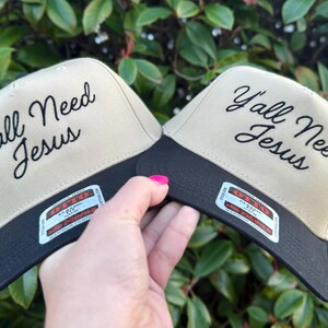 May include: Two beige and black baseball caps with the text "Y'all Need Jesus" embroidered in black on the front of each cap.