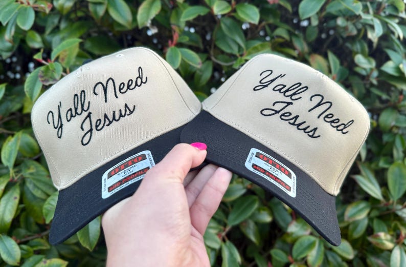 May include: Two beige and black baseball caps with the text "Y'all Need Jesus" embroidered in black on the front of each cap.