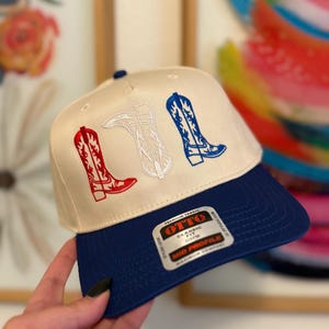 May include: A white and blue baseball cap with a red, white, and blue embroidered design of cowboy boots. The cap has a blue brim and a white label with the text "OTTO CLASSIC FIT OSFM MID PROFILE".