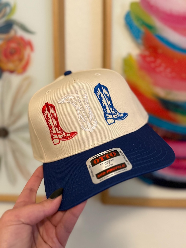 May include: A white and blue baseball cap with a red, white, and blue embroidered design of cowboy boots. The cap has a blue brim and a white label with the text "OTTO CLASSIC FIT OSFM MID PROFILE".
