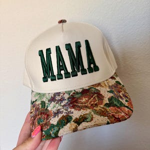 Puede incluir: Gorra de b&eacute;isbol color crema con la palabra "MAMA" bordada en letras verdes oscuras. La visera presenta un dise&ntilde;o de tapiz floral en tonos verdes, rojos y marrones. Un peque&ntilde;o bot&oacute;n decorativo est&aacute; en la parte superior.