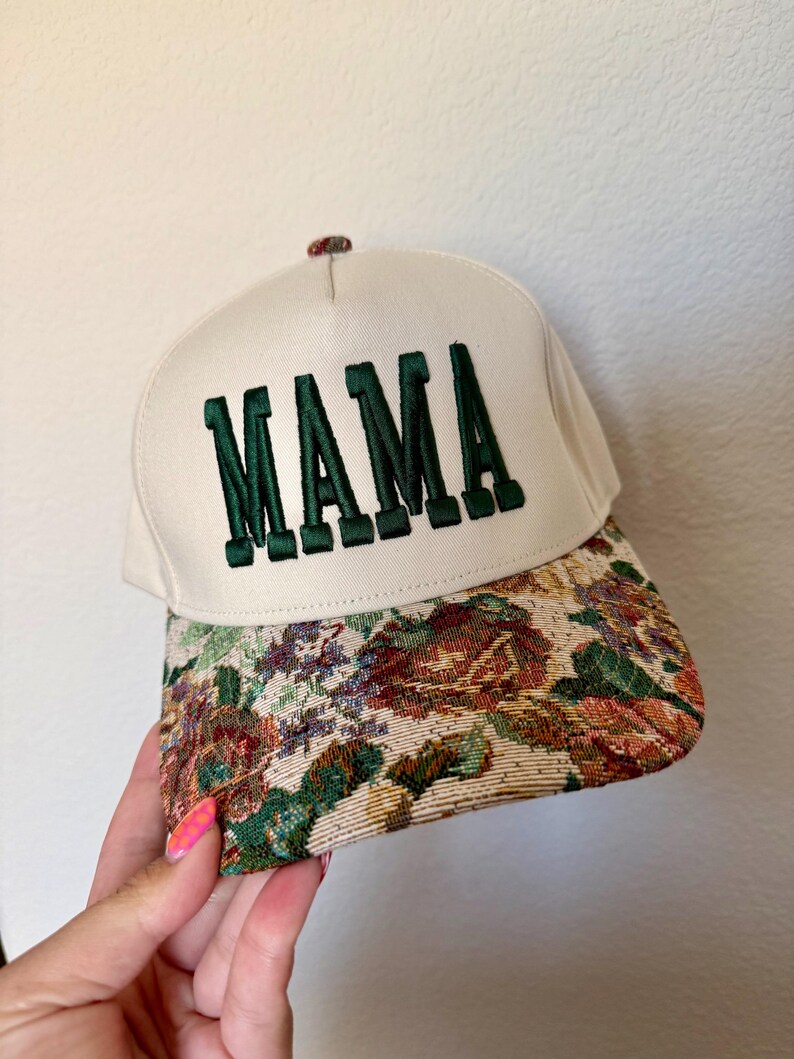 Puede incluir: Gorra de b&eacute;isbol color crema con la palabra "MAMA" bordada en letras verdes oscuras. La visera presenta un dise&ntilde;o de tapiz floral en tonos verdes, rojos y marrones. Un peque&ntilde;o bot&oacute;n decorativo est&aacute; en la parte superior.