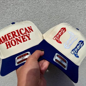May include: Two trucker hats with a blue and white color scheme. One hat has the text "AMERICAN HONEY" embroidered in red on the front. The other hat has a white cowboy boot design with red and white accents.