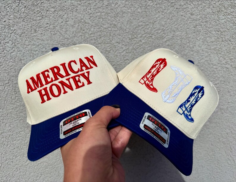 May include: Two trucker hats with a blue and white color scheme. One hat has the text "AMERICAN HONEY" embroidered in red on the front. The other hat has a white cowboy boot design with red and white accents.